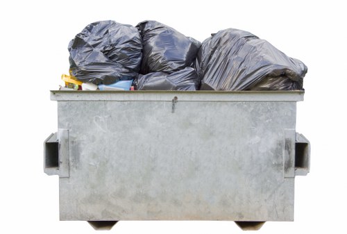 A large, rectangular metal skip bin positioned outdoors on a flat surface, filled with multiple tightly packed black rubbish bags made of thick plastic. The bags have a slightly crinkled texture and are arranged in an uneven stack, with some leaning on each other. The skip's surface appears weathered with visible scratches and minor rust, and it features reinforced handles on each side. The background is plain and neutral, suggesting the bin is situated in a driveway or yard, possibly in the Brent Cross area, as part of a rubbish removal or clearance service by House Clearance Brent Cross. Natural daylight illuminates the scene, highlighting the metallic finish of the bin and the dark tones of the rubbish bags, underscoring the focus on waste collection and disposal processes typical of local waste management efforts.